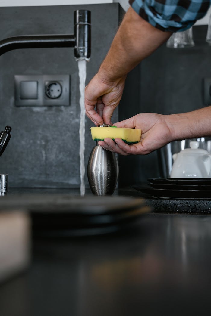 Close-up of a person cleaning dishes with a sponge under running water in a modern kitchen setting.